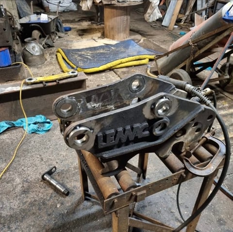 Black metal hydraulic excavator bucket attachment with cylinders on workshop workbench, surrounded by tools and equipment