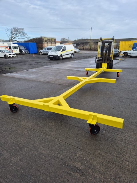 Yellow forklift tow bar attachment in an industrial yard with parked vans and construction equipment